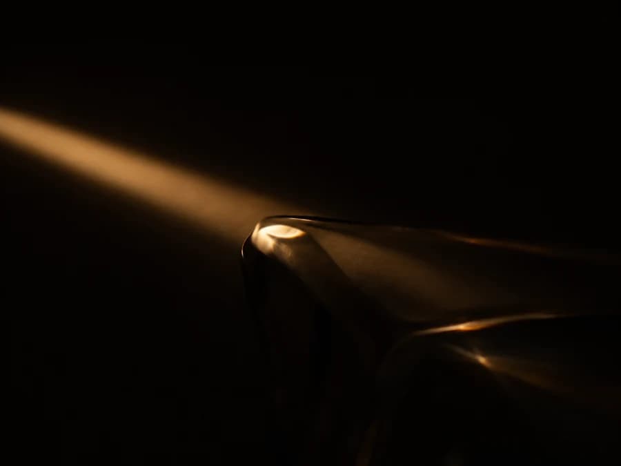A thin beam of warm light cuts through darkness, revealing the edge of a glass form